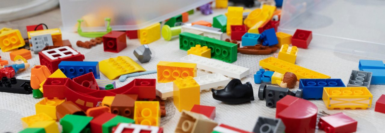 Vista de cima de brinquedos coloridos espalhados no chão, incluindo blocos de montar, carrinhos, e peças de encaixe, organizados de forma desordenada sobre uma superfície clara.